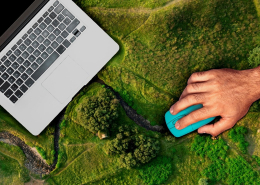 A hand with a mouse operates a laptop for monitoring, reporting, and verification, which is placed on a landscape viewed from above.