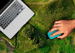 A hand with a mouse operates a laptop for monitoring, reporting, and verification, which is placed on a landscape viewed from above.